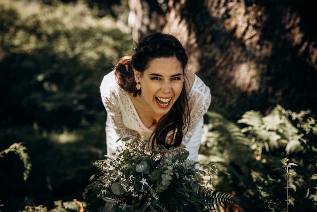 une mariée se tient dans la nature avec son bouquet de mariée très végétal dans un dégradé de vert et blanc avec des fleurs délicates et de différentes variétés de feuillage comme de la fougère et de l'eucalyptus et éclate de rire