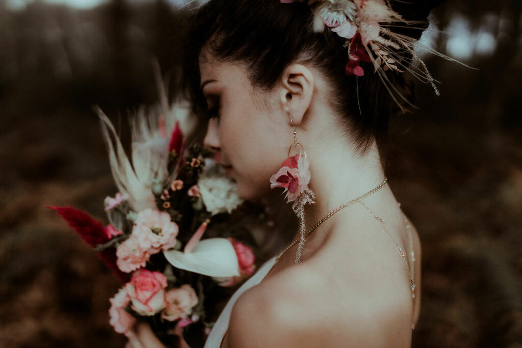 une mariée de profil ferme les yeux tenant dans les mains son bouquet de mariée graphique composé d'un anthurium, de roses, de lisianthus et de plumes de paon blanches, portant des boucles d'oreilles et un peigne fleuris assortis