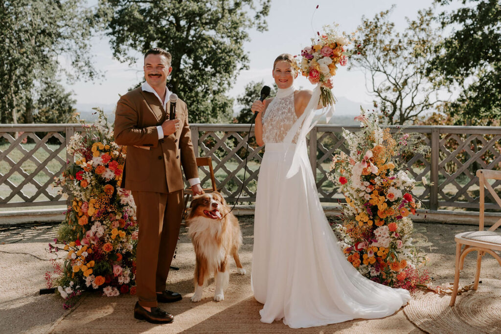 couple de mariés et leur chien, installés devant la décoration florale composé de deux totems de fleurs très champêtres de couleurs très vivantes, chatoyantes de couleurs chaudes lors de leur cérémonie en extérieur au Domaine de Larraldia à Villefranque au Pays basque