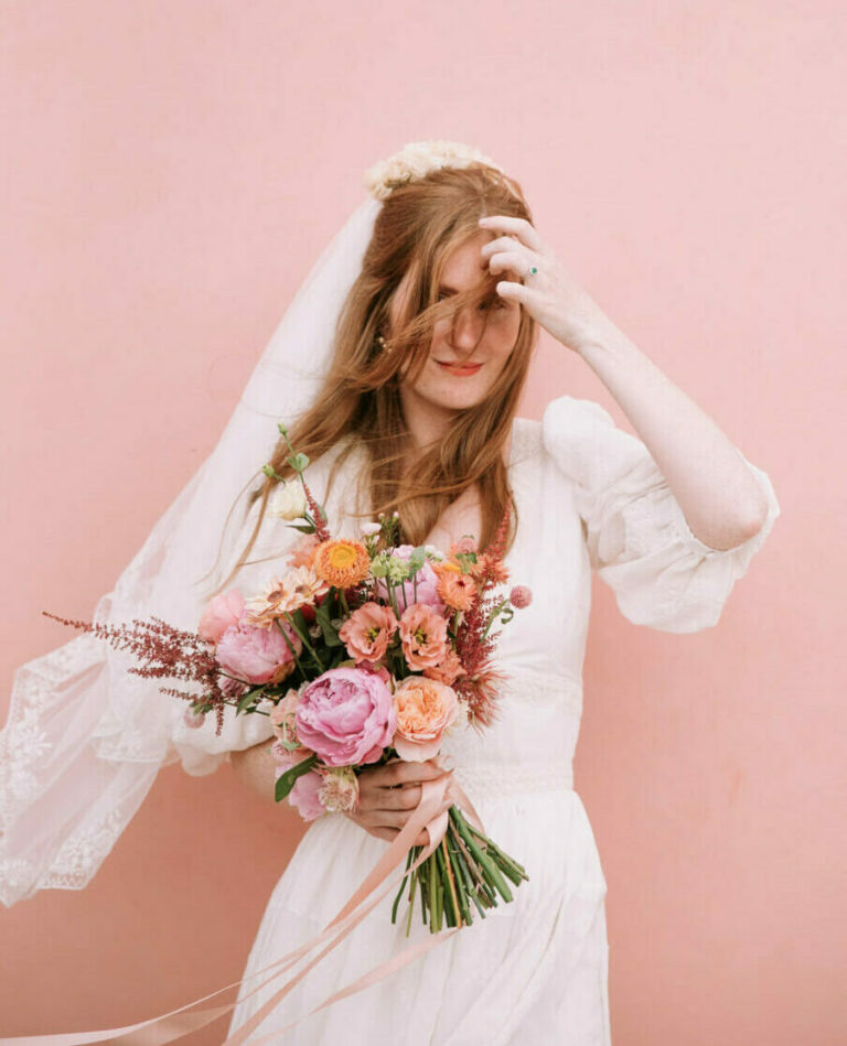 la mariée pose devant un mur rose en tenant son bouquet et se touche les cheveux qui flottent dans le vent