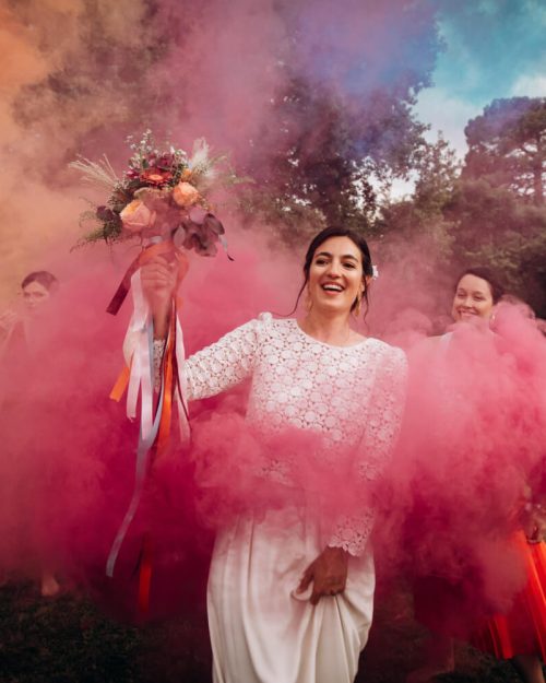une mariée souriante est entourée de ses demoiselles d'honneur lors d'une seance photo de groupe avec des fumigènes de couleurs multicolores et porte son bouquet de mariée dans la main droite en hauteur après le jeu du ruban